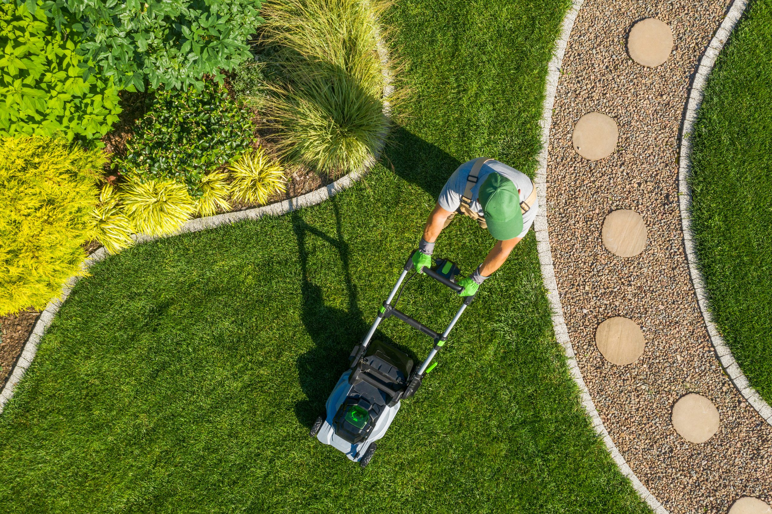 A person mows a neatly landscaped lawn, surrounded by various plants and stones