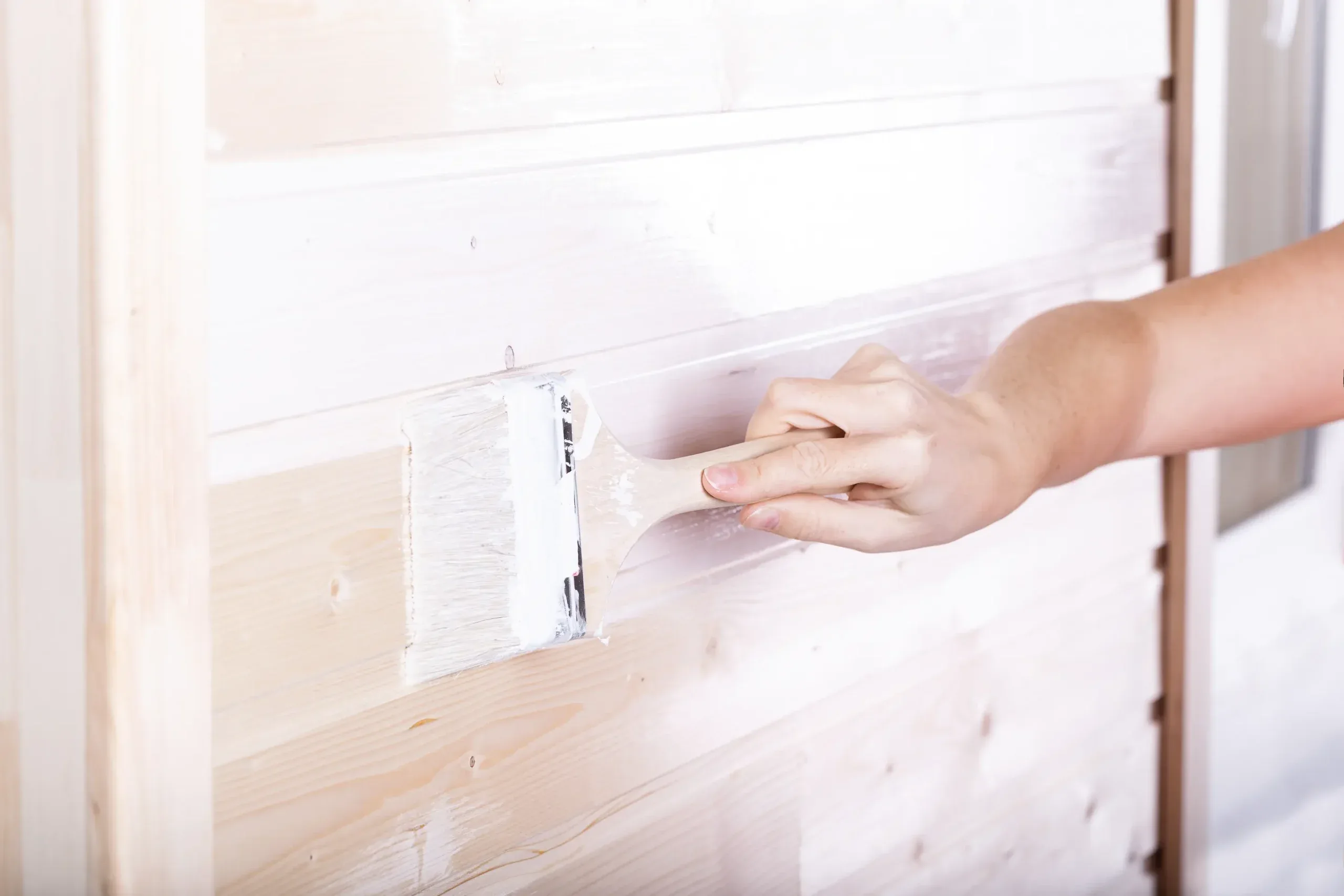 A hand applies paint to a wooden wall with a brush