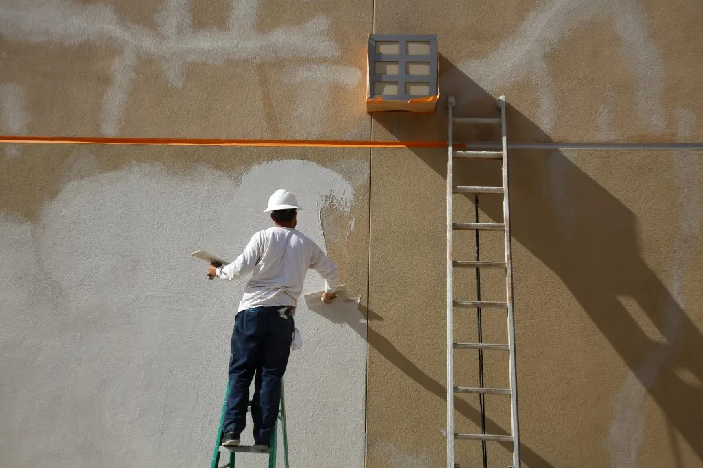 A person in a white hard hat paints a wall while standing on a ladder