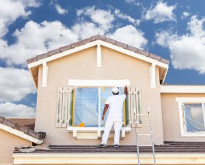 A person in white overalls paints a window on a house under a bright sky