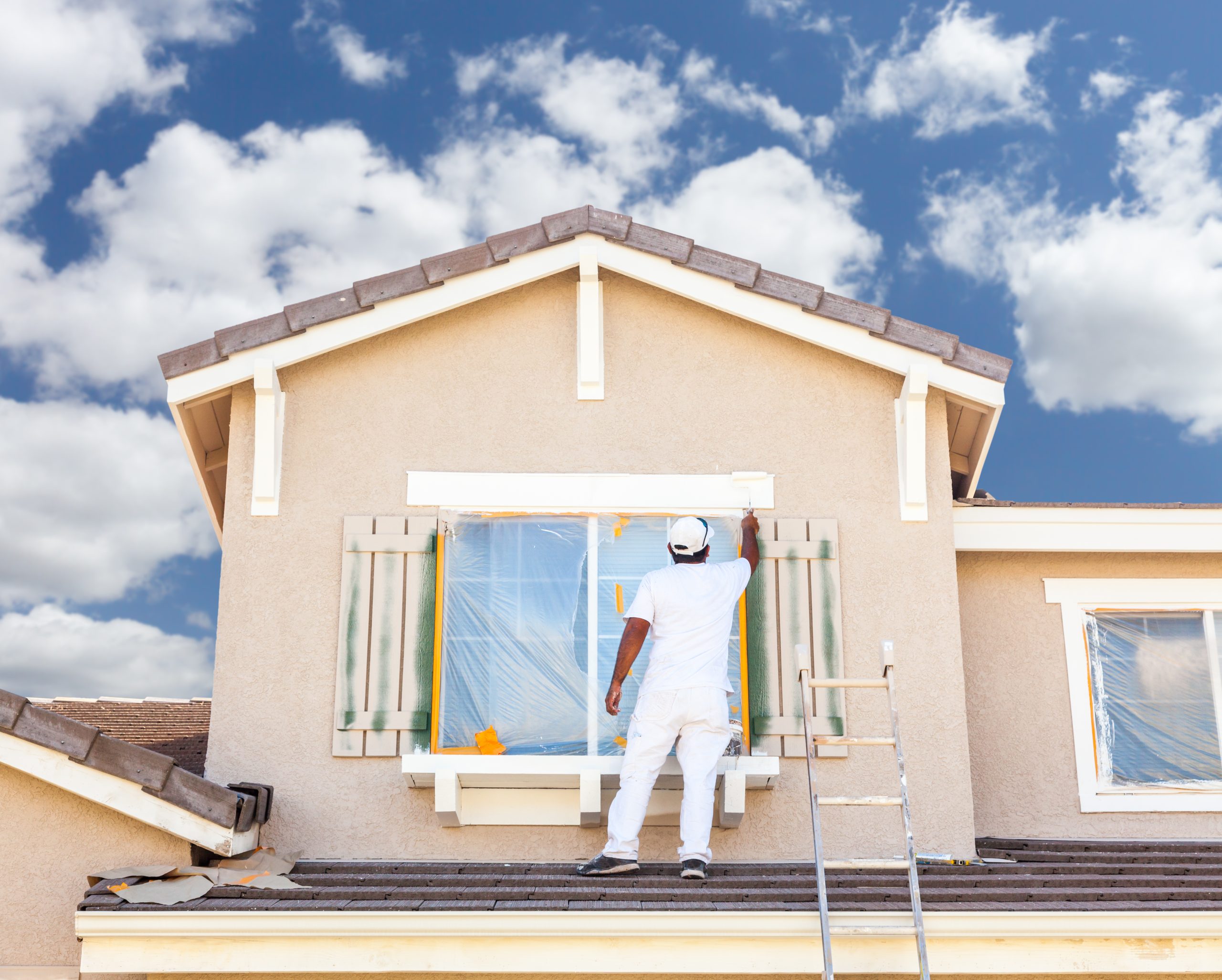 A person in white overalls paints a window on a house under a bright sky
